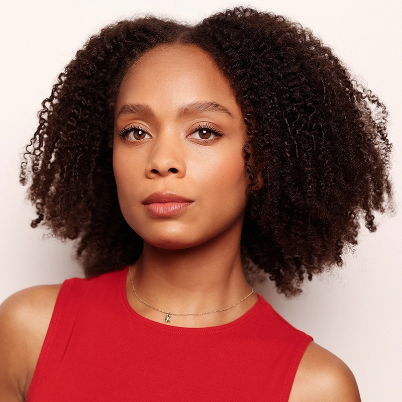 Talia Robinson, a young woman with medium brown skin and natural, voluminous curly hair, wears a sleeveless red top and a delicate gold necklace. She looks confidently at the camera against a light, neutral background.