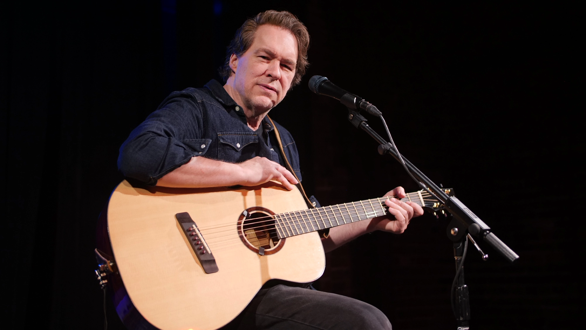 A man in a dark denim shirt sits on stage, playing an acoustic guitar and looking toward the camera, evoking the introspective style of Simon. He is mid-performance, with a microphone in front of him against a dark background.