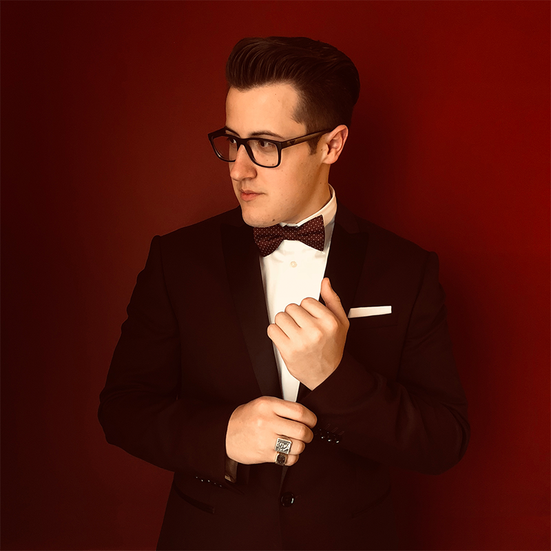 Wyatt Michael, in a black tuxedo, white shirt, and bow tie, stands against a deep red background. He wears glasses, a ring, and a white pocket square, looking to the side with one hand adjusting his jacket.