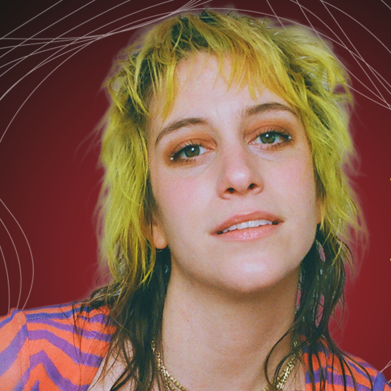 A person with shaggy, yellow-blonde hair and dark roots gazes softly at the camera. They wear a colorful, striped top with purple and orange tones, gold necklaces, and subtle makeup. The background is deep red with white, swirling line patterns.