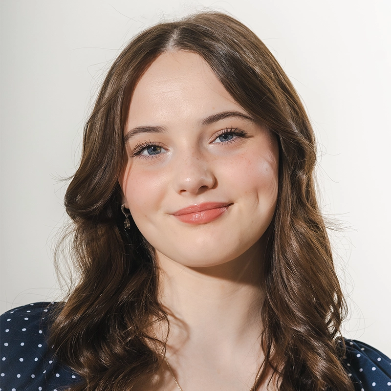 A young woman with fair skin, long wavy brown hair, and blue eyes smiles softly. Ireyssa Cardona wears a navy blue polka-dot top and natural makeup. The background is plain white, and the lighting is bright and even across her face.