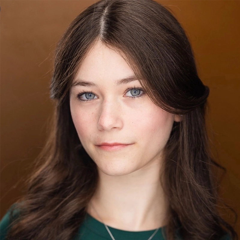 Sara-Louise Oakes, a young woman with long wavy brown hair and blue eyes, looks at the camera with a neutral expression. She wears a dark green top against a warm brown background, with soft lighting highlighting her clear skin and delicate features.