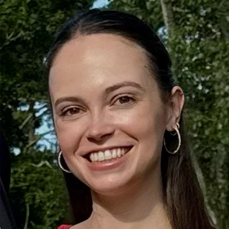 Abby Wolanin, with straight dark brown hair pulled back, wearing hoop earrings and a red top, smiles at the camera. The background features green trees and sunlight, suggesting an outdoor setting.