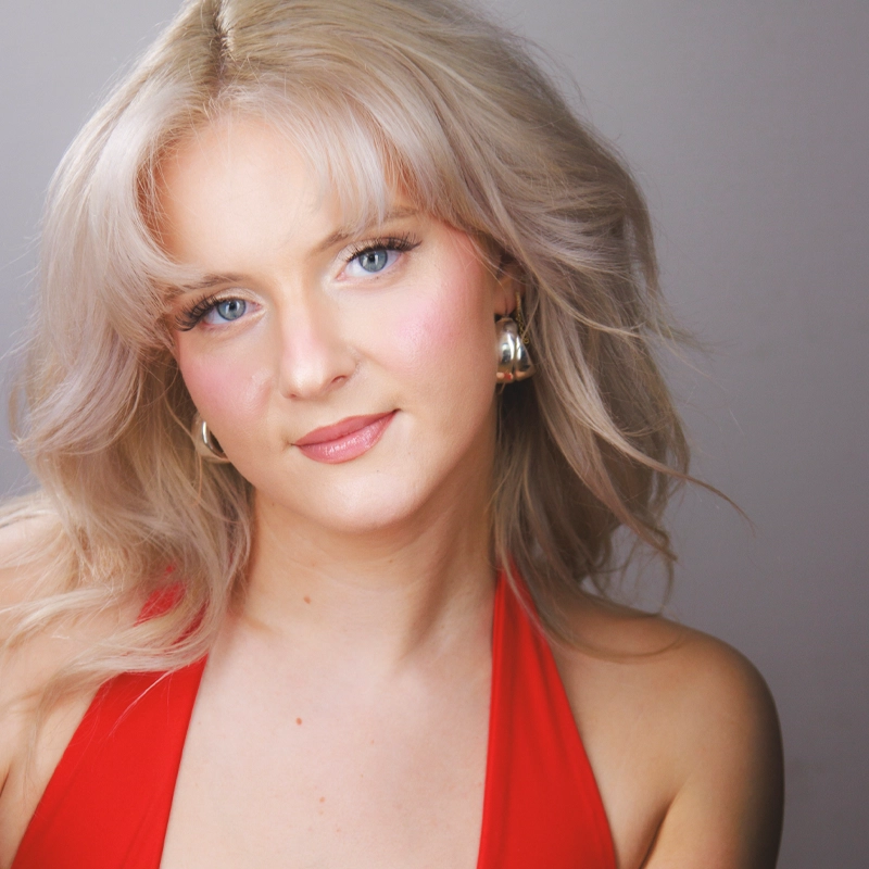 Katherine Abel, a young woman with wavy platinum blonde hair and blue eyes, poses in front of a plain gray background. She smiles gently at the camera, wearing a sleeveless red top, silver hoop earrings, and soft pink makeup.