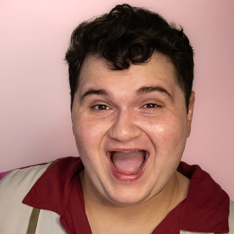 Aidan Michael Kelly, with short, dark, curly hair, smiles widely with their mouth open. Wearing a maroon and beige shirt against a soft pink background, their expression appears happy and enthusiastic.