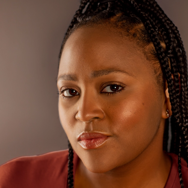A woman with long, braided hair looks directly at the camera with a confident expression. She wears subtle makeup and a reddish-brown top. The background is a soft, neutral gray, and warm lighting highlights her smooth skin and features.