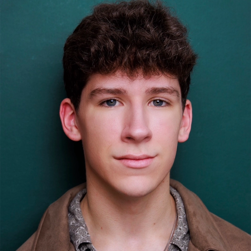 Young man with short, curly brown hair and clear skin facing forward against a dark green background. He wears a brown jacket over a shirt with a subtle pattern. With blue eyes, a slight smile, and an overall calm, neutral expression, he closely resembles Killian Allgeier.