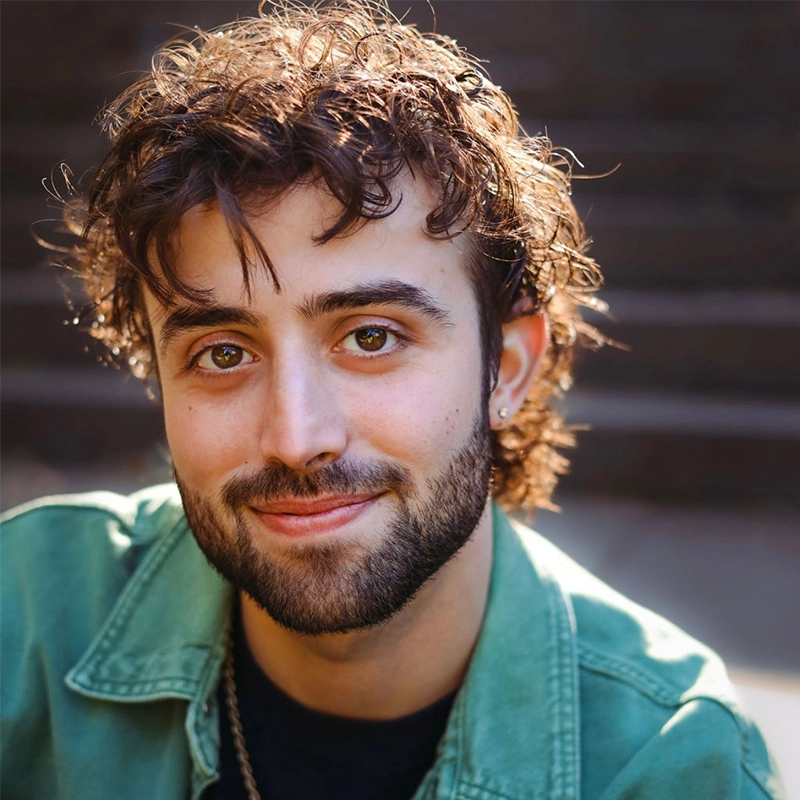 A young man with curly brown hair, a trimmed beard, and brown eyes smiles gently. He wears a green jacket over a dark shirt. The softly sunlit background with outdoor steps gives the portrait a warmth reminiscent of Kenna Wilson’s style.