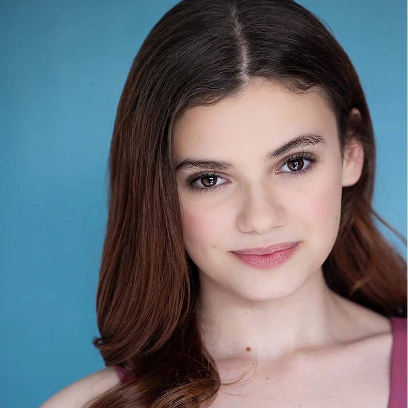 A young woman with long brown hair and brown eyes smiles softly at the camera. Katie Krasovec has fair skin, subtle makeup, and wears a mauve top. The solid light blue background gives the portrait a bright and fresh appearance.