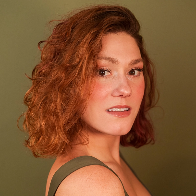Katie Yeomans, with fair skin and shoulder-length, wavy auburn hair, poses against an olive green background. She wears a sleeveless green top and looks confidently at the camera with a soft smile and natural makeup.