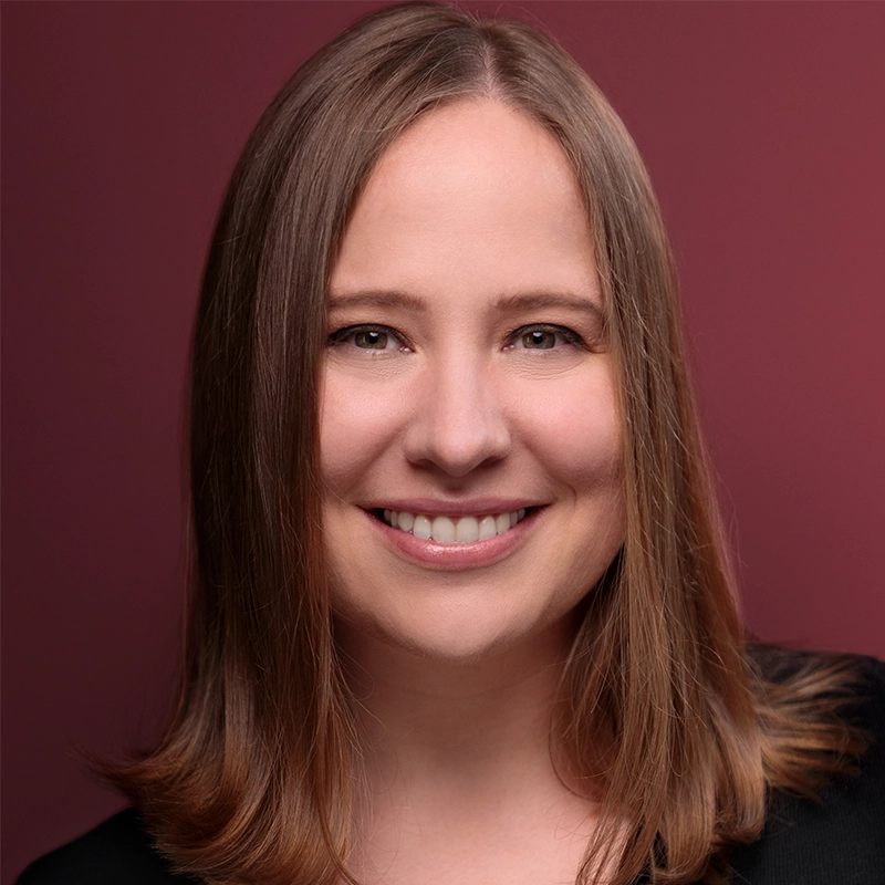 Arlee Chadwick, with straight, light brown hair parted slightly to the side, smiles at the camera. She is wearing a black top against a solid maroon background. Soft lighting highlights her friendly expression.
