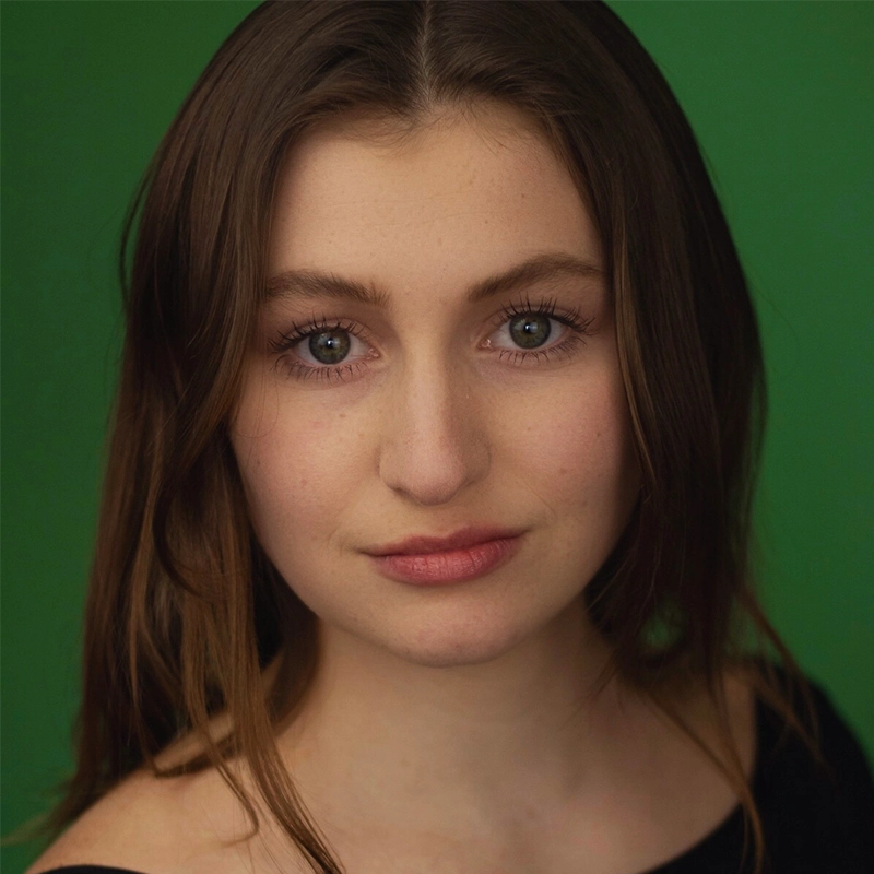 Aidan Michael Kelly captures a young woman with long brown hair and blue-green eyes gazing into the camera. She has fair skin, natural makeup, and wears an off-the-shoulder black top against a solid green background.