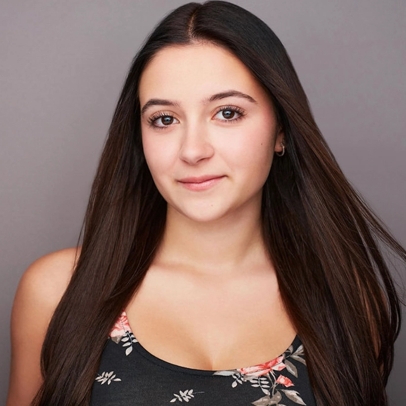 Carissa Ferguson, a young woman with long straight brown hair and a slight smile, faces the camera against a plain gray background. She wears a black tank top with a pink and white floral pattern, her natural makeup highlighting her calm and confident expression.