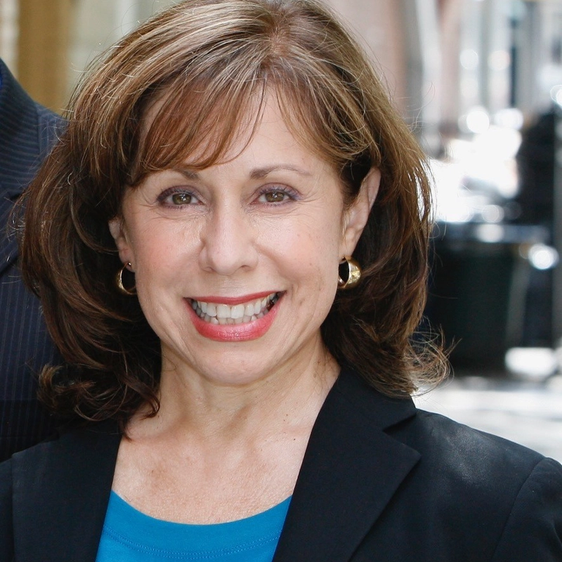 Lynnette Barkley, a woman with shoulder-length brown hair and bangs, smiles at the camera. She is wearing gold hoop earrings, a blue top, and a black blazer against a softly blurred outdoor urban backdrop with buildings and lights.