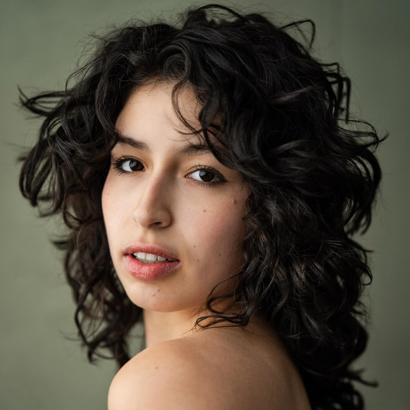 Gabriela Barrett, a young woman with wavy, dark hair, looks over her shoulder at the camera. She has a natural expression and minimal makeup, with soft lighting highlighting her clear skin and brown eyes against a muted olive green background.