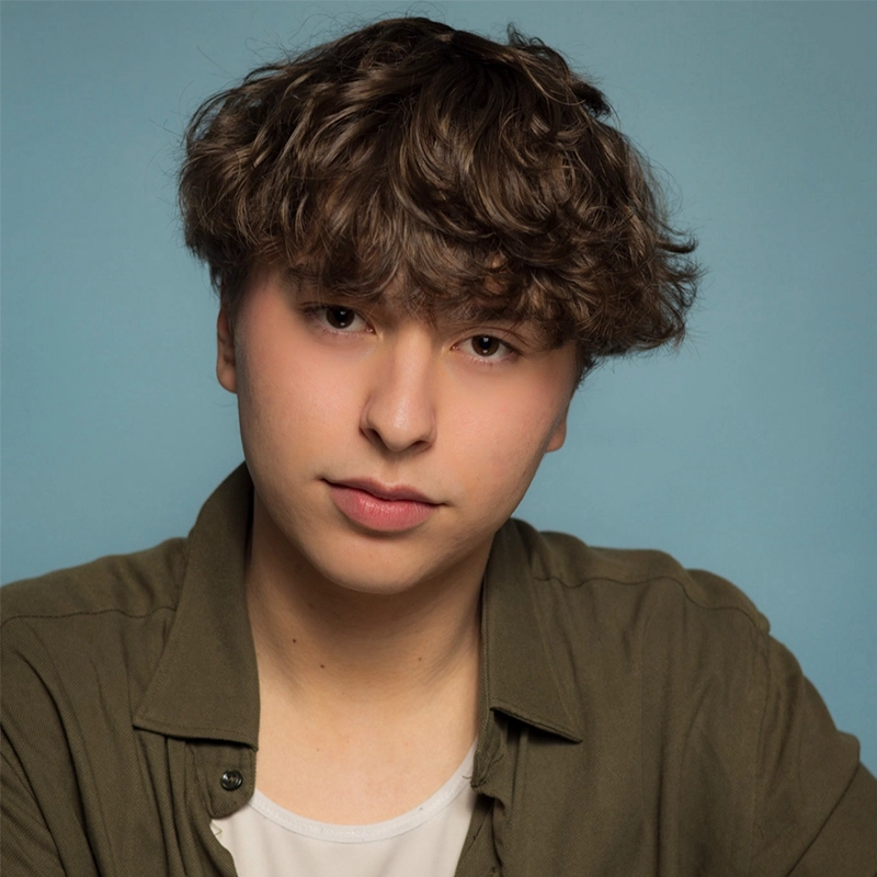 A young person with wavy brown hair and fair skin looks at the camera. They wear an olive-green button-up shirt over a white top, set against a light blue background. Their neutral, calm expression resembles portraits of Killian Allgeier.