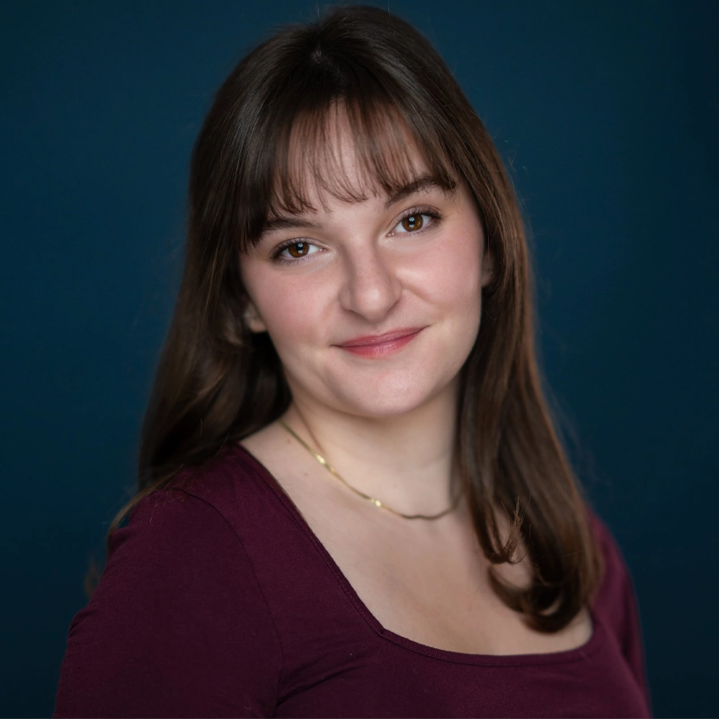 A young woman, Lauren Biedron, with straight brown hair and bangs smiles slightly. She wears a burgundy top and a thin gold necklace. The background is a solid dark blue. The lighting is soft, highlighting her face and shoulders.