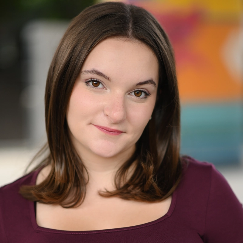 Lauren Biedron, a young woman with straight brown hair and fair skin, smiles slightly in a maroon top with a square neckline. The background is softly blurred with vibrant colors—orange, pink, and teal shapes.