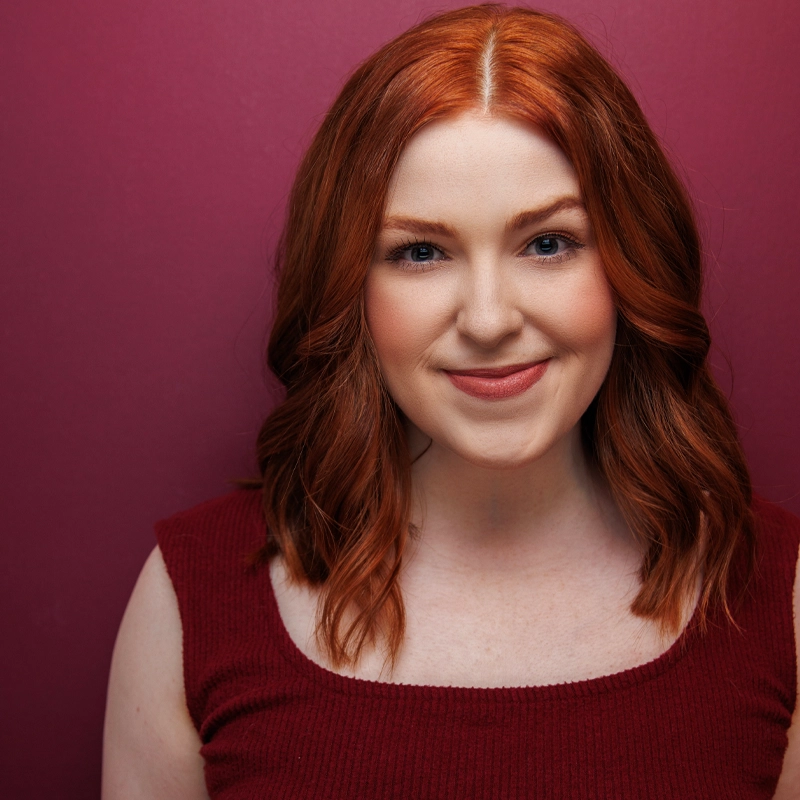 Maddy Mae Billings, a woman with fair skin and shoulder-length wavy red hair, smiles gently. She wears a sleeveless, dark red top and stands against a solid burgundy background, the lighting highlighting her hair and features.