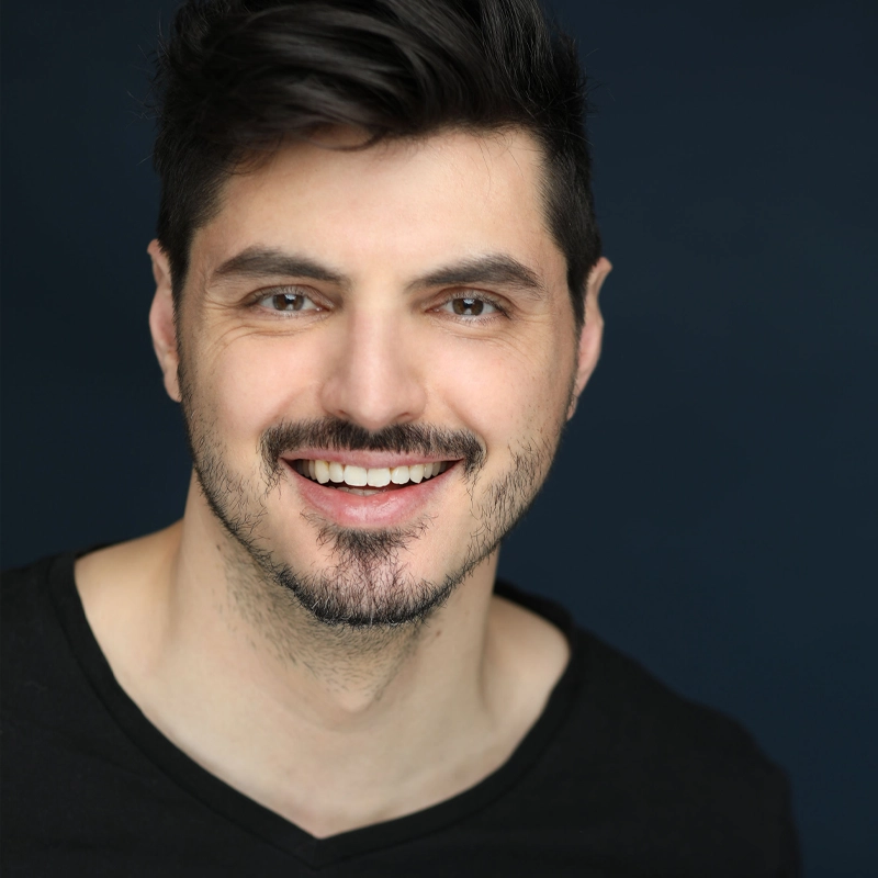 Chris Boccard, a man with short dark hair and facial hair, smiles warmly at the camera. He wears a black V-neck shirt and stands against a dark blue background, the lighting highlighting his friendly and approachable demeanor.