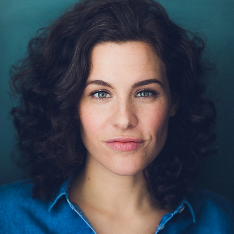 Sarah Bockel, a woman with fair skin, blue-green eyes, and dark curly hair, gazes at the camera with a slight smile. She wears a blue denim shirt and poses before a dark teal background under soft, even lighting.