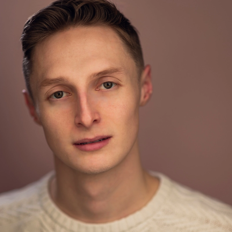 A young man with short, light brown hair and fair skin gazes softly at the camera. Brett Boline wears a cream-colored knit sweater. The background is blurred and warm-toned, creating a gentle, neutral setting with soft, even lighting.