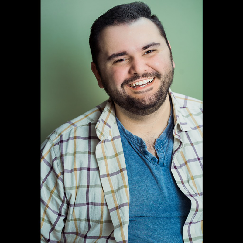 Bradley Ford Betros, a man with short dark hair and a full beard, smiles warmly at the camera. He wears a light plaid shirt over a blue henley. The soft green background is framed by black bars on each side.