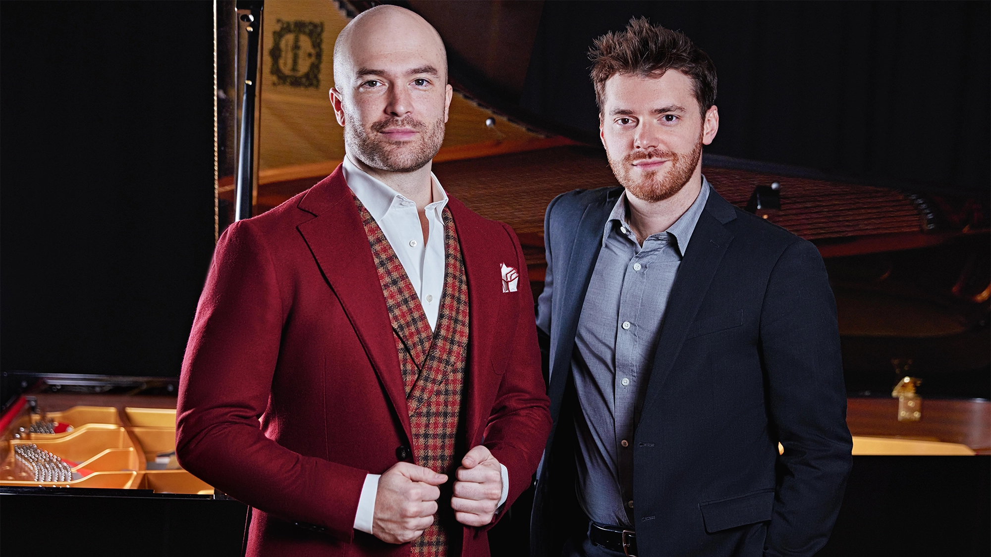 Brancy & Dugan site image 25 Two men stand in front of an open grand piano. The man on the left wears a red suit with a patterned vest, white shirt, and pocket square, with his hands on his jacket. The man on the right wears a dark suit and shirt. Both look confidently at the camera.