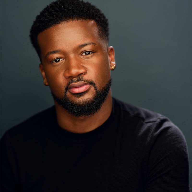 Dewight Braxton Jr., with short curly hair and a trimmed beard, looks at the camera with a neutral expression. He wears small gold earrings and a black crew-neck shirt against a dark, softly blurred background, his face highlighted by the lighting.