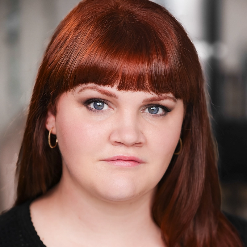 A woman with straight, reddish-brown hair and blunt bangs looks directly at the camera. She has fair skin, blue eyes, gold hoop earrings, and wears a neutral expression with subtle makeup. The background is softly blurred in neutral tones.