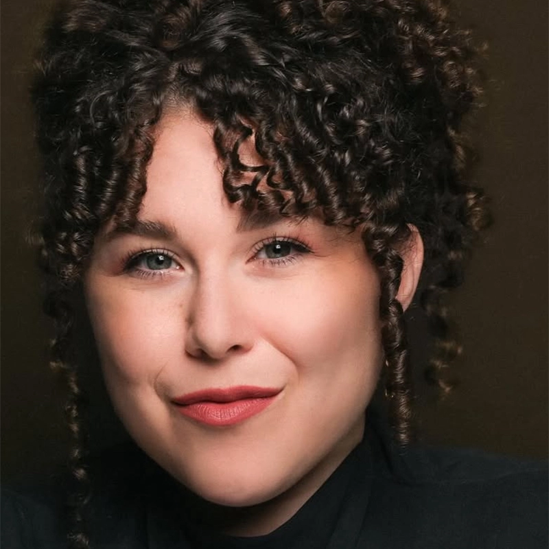 Kenna Wilson, with fair skin and curly dark brown hair, smiles softly. Her blue eyes, full eyebrows, and pink lipstick stand out as spiral curls frame her face. She wears a dark top and poses against a plain, dark background.