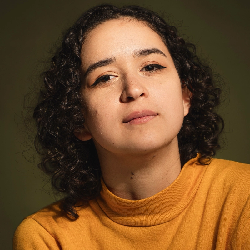 Lauren Brown, with short, dark, curly hair and winged eyeliner, looks confidently at the camera. She wears a mustard yellow turtleneck sweater against an olive green background, the soft lighting highlighting her face and expression.