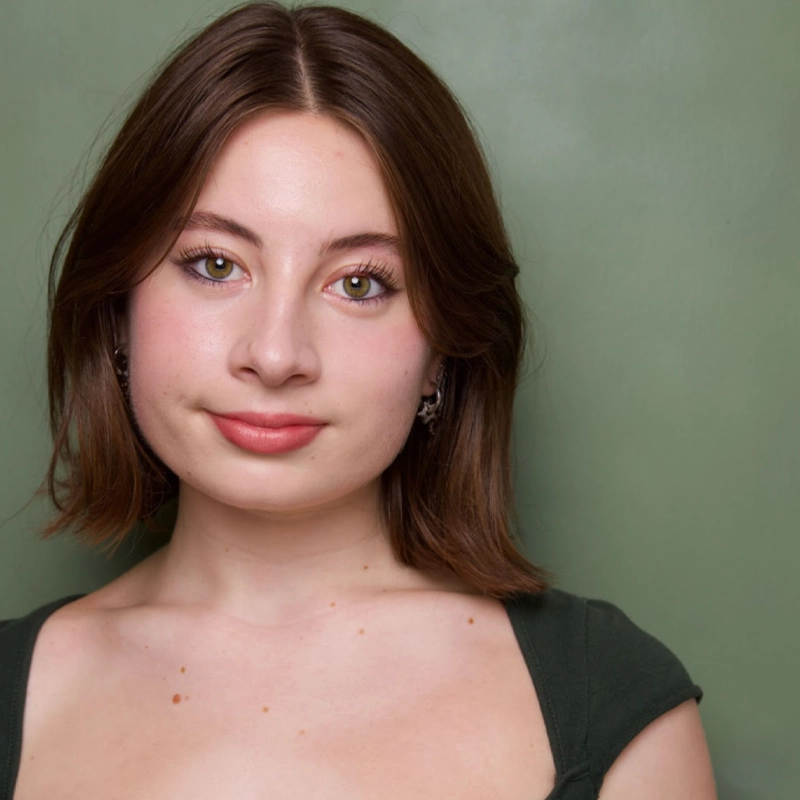 A young woman with fair skin and shoulder-length brown hair stands against a muted green background. Daphne Bruhmuller has light hazel eyes, subtle makeup, and wears a dark green top, facing the camera with a gentle, closed-mouth smile.