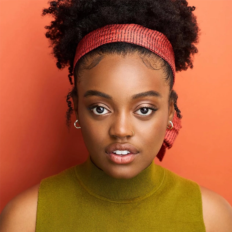 A young woman with dark curly hair in a red headband, wearing small hoop earrings and a sleeveless olive-green turtleneck, looks directly at the camera against an orange background. She has a natural makeup look and a neutral expression.