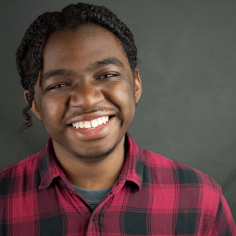 Myles Carter, a young man with medium-dark skin and tightly braided hair, smiles warmly at the camera. He wears a red and black plaid shirt over a dark undershirt, with the plain gray background highlighting his cheerful expression.