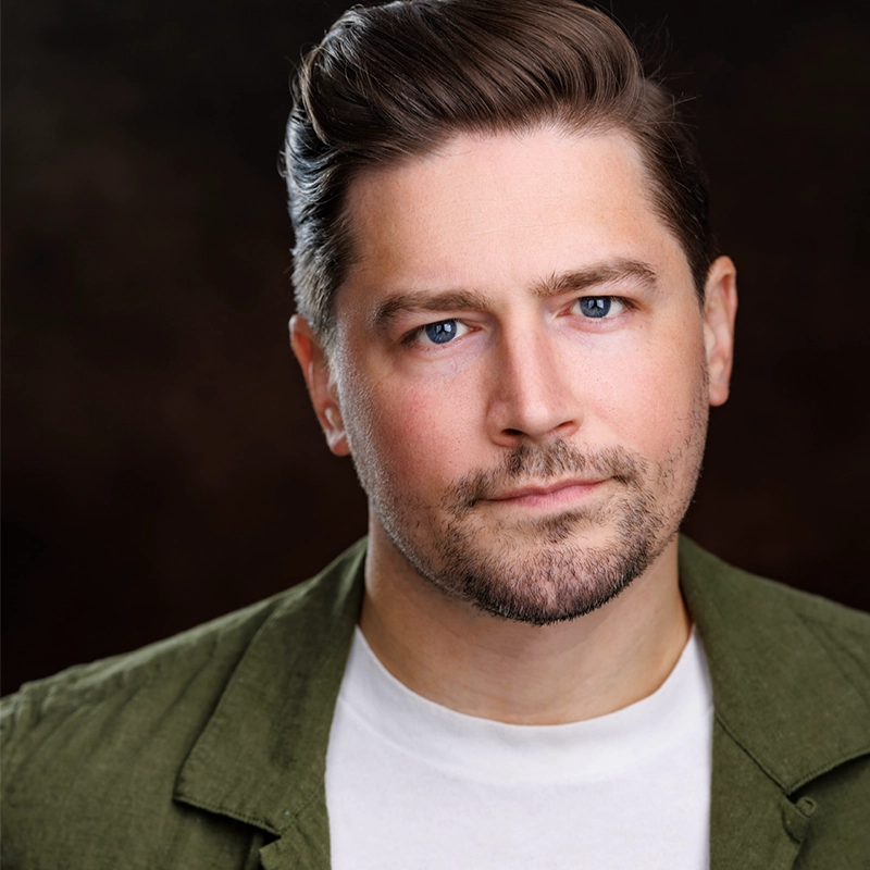 A man with short, neatly styled brown hair and light stubble, wearing a white t-shirt and an olive green jacket, looks directly at the camera with a neutral expression against a dark, blurred background by Katie Yeomans.