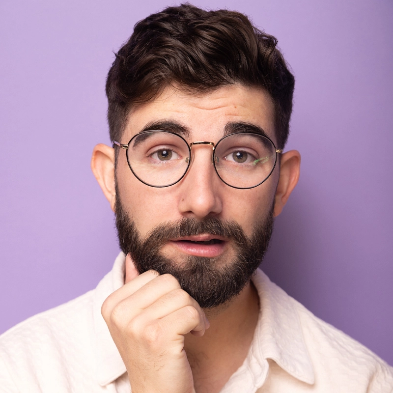 Peyton Chance, a young man with short brown hair and a full beard, gazes thoughtfully at the camera. He wears round glasses and a white collared shirt, his hand resting on his chin against a plain light purple background.