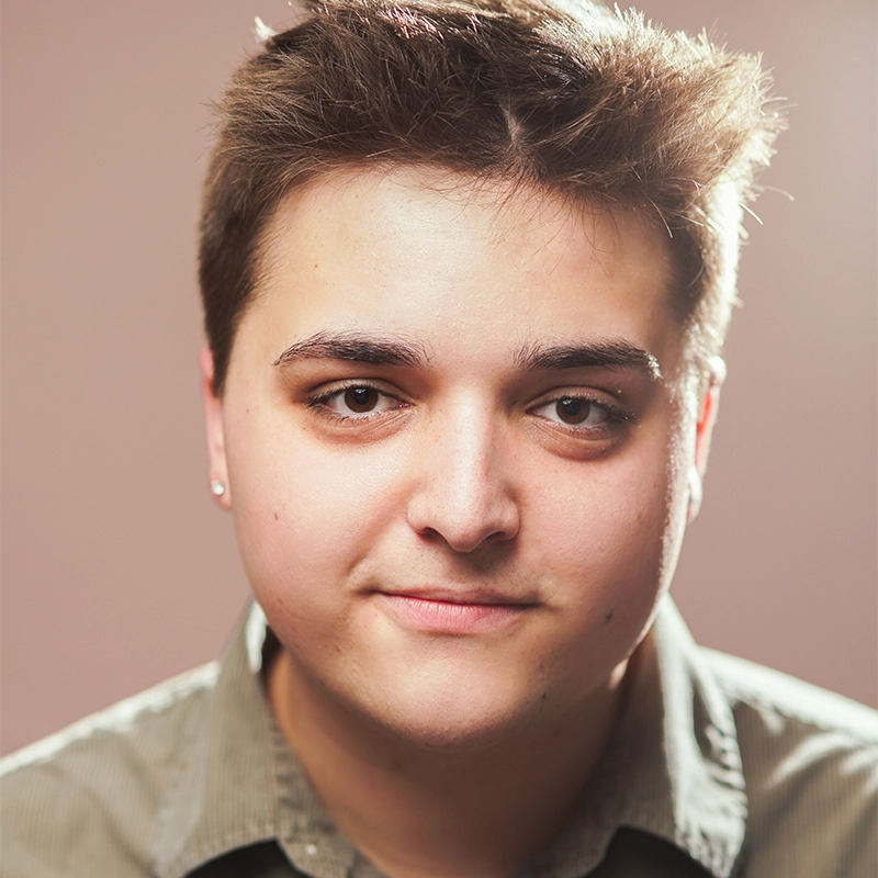 Charles Pelletier, with short brown hair and light skin, looks directly at the camera with a slight smile. Wearing a collared shirt and a small earring, he stands against a softly blurred, neutral background with soft, even lighting.