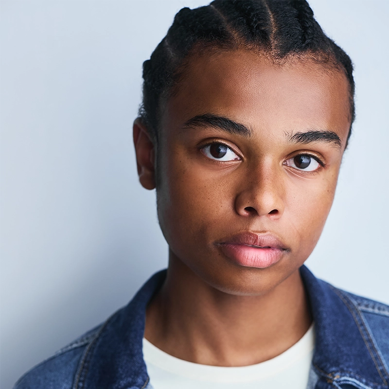 A young person with medium brown skin and braided hair looks directly at the camera with a neutral expression. Lyla Karekinian wears a white shirt and a denim jacket, standing against a plain, light background that highlights her calm demeanor.