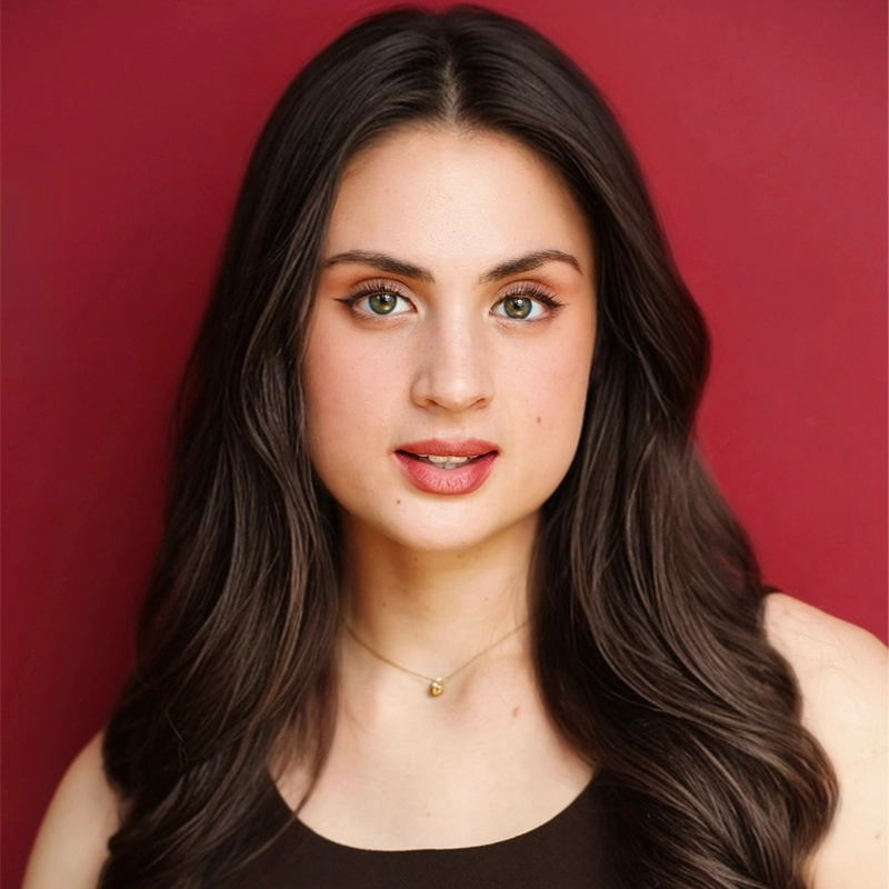 Katie Krasovec, a young woman with long, wavy dark brown hair and hazel eyes, poses in front of a solid red background. She wears a sleeveless black top and delicate gold necklace, looking directly at the camera with a calm, neutral expression.