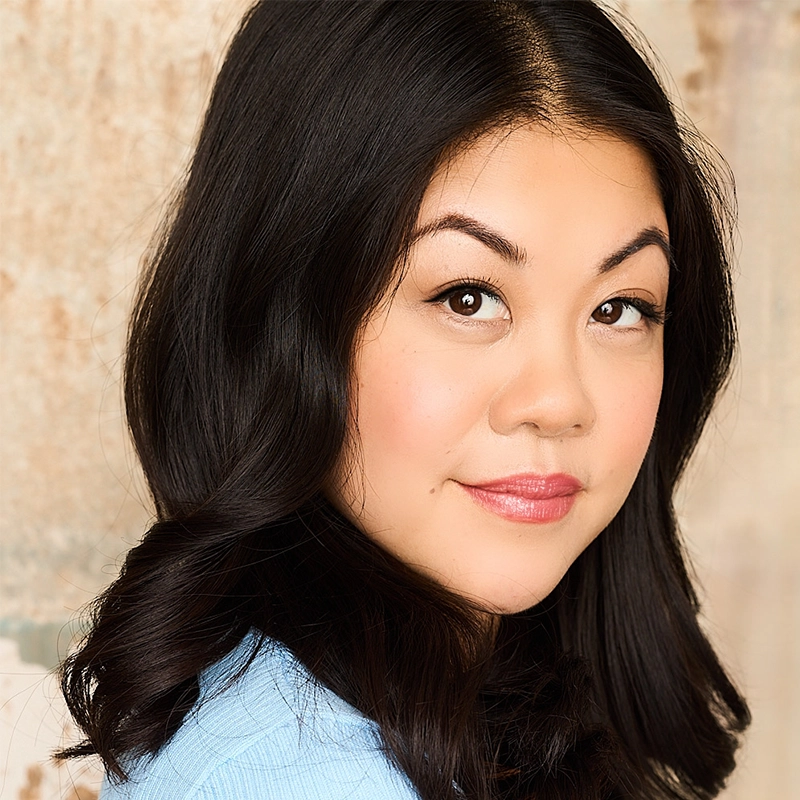 Charlotte Mary Wen, with long, wavy black hair and smooth fair skin, gazes at the camera. She has defined eyebrows, winged eyeliner, light blush, and pink lipstick. She wears a light blue top against a softly blurred neutral background.