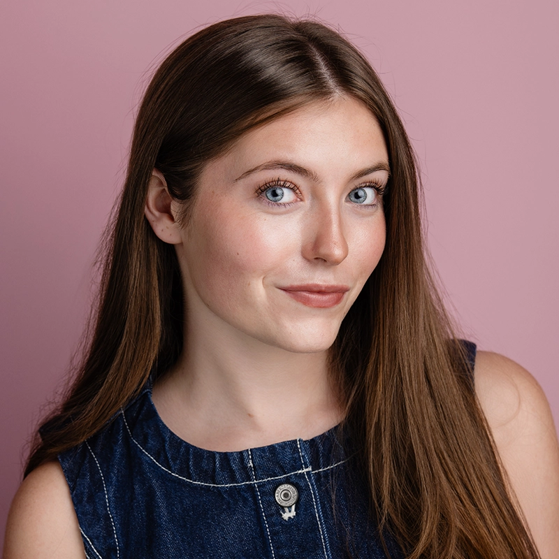 Carissa Ferguson, a young woman with long straight brown hair and light eyes, smiles gently. She wears a dark denim sleeveless top with white stitching and a round button at the collar, against a solid pink background as she faces slightly left of the camera.