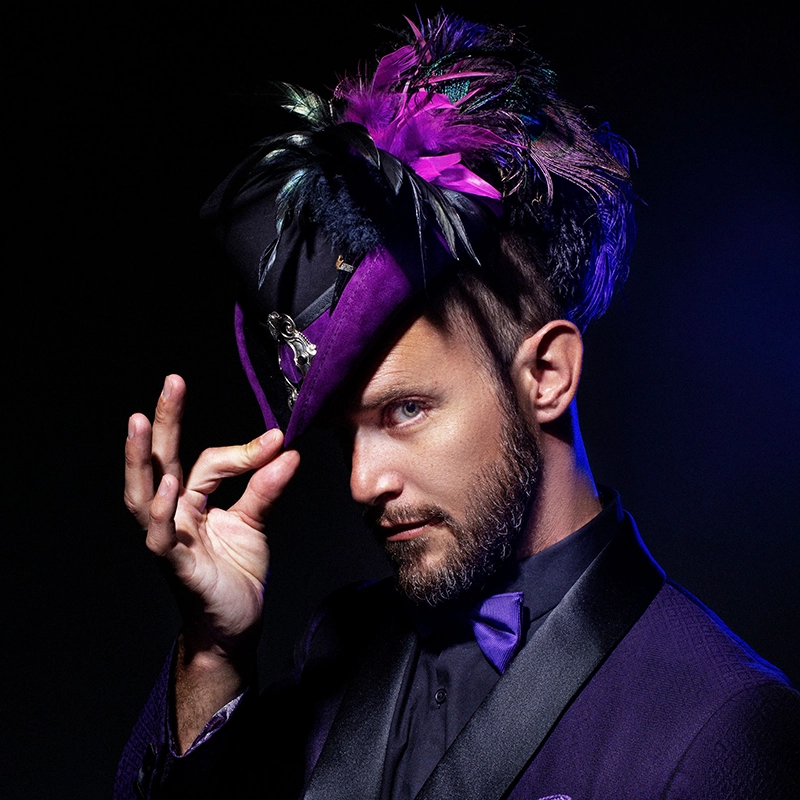 A man with a trimmed beard tips a purple hat adorned with black and purple feathers, channeling Latrice Royale elegance. He wears a matching purple suit, shirt, and bow tie. Dramatic lighting highlights his face against a dark background.