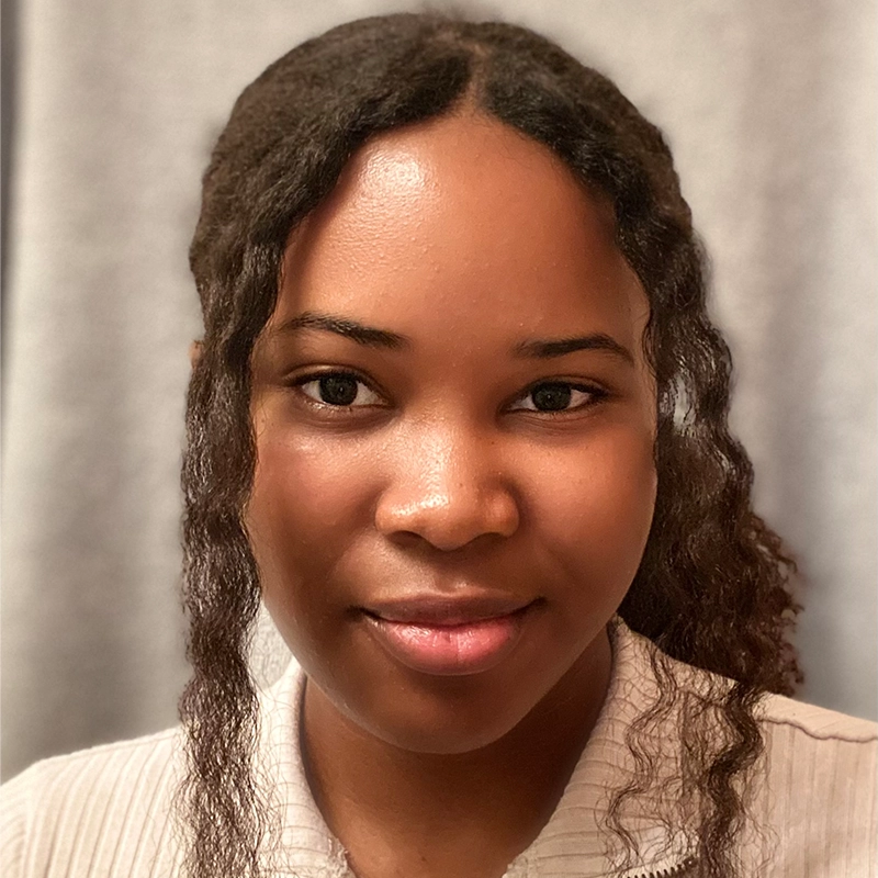 A young woman with medium-dark skin and long, wavy dark hair, parted in the middle. Katie Krasovec is wearing a light-colored, textured top and smiling softly. The background is a plain, light gray fabric or curtain.