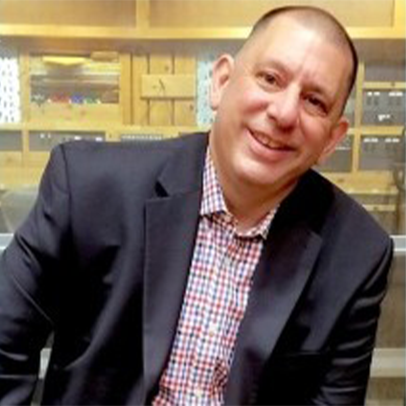 A smiling man with short hair, identified as Chuck Granata, wears a dark blazer over a red and white checked shirt, sitting slightly angled in front of wooden shelves filled with various items in a warmly lit indoor setting.