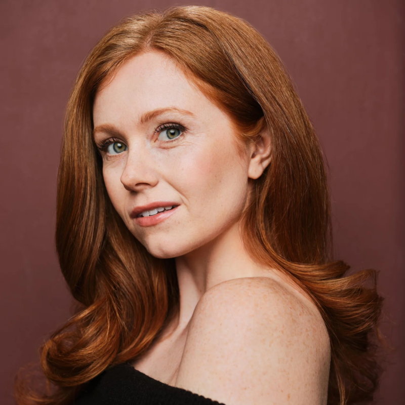 A woman with long, wavy red hair poses in front of a muted mauve background. She has light skin with freckles, green eyes, and is wearing a black off-the-shoulder top. She gazes softly at the camera, slightly smiling.
