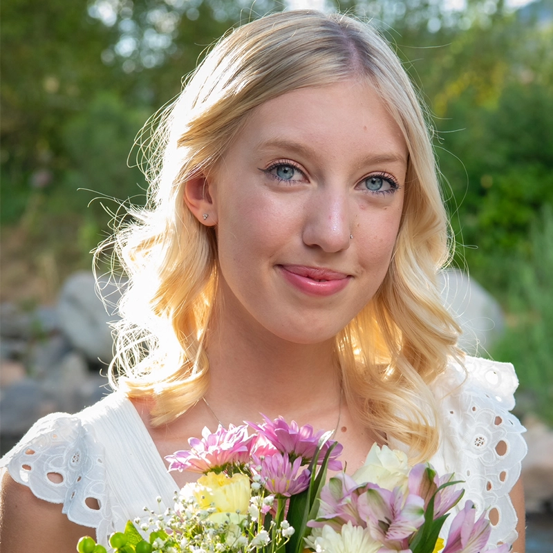 A young woman with light blonde hair and blue eyes smiles gently, holding a bouquet of colorful flowers. Carissa Ferguson wears a white top with eyelet details as sunlight softly illuminates her hair against a blurred green background.