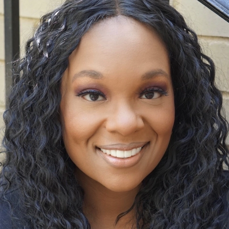 Carrie Compere, with long, wavy black hair, smiles warmly at the camera. She has smooth brown skin, wears natural makeup with subtle eye shadow and lipstick, and is pictured outdoors by a beige brick wall and black railing.