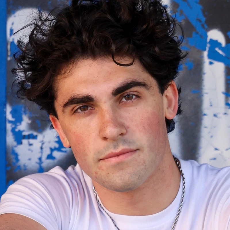 Aiden Connolly, a young man with curly dark hair and bushy eyebrows, wears a white T-shirt and silver chain necklace. He looks directly at the camera with a neutral expression against a backdrop of blue and white graffiti.