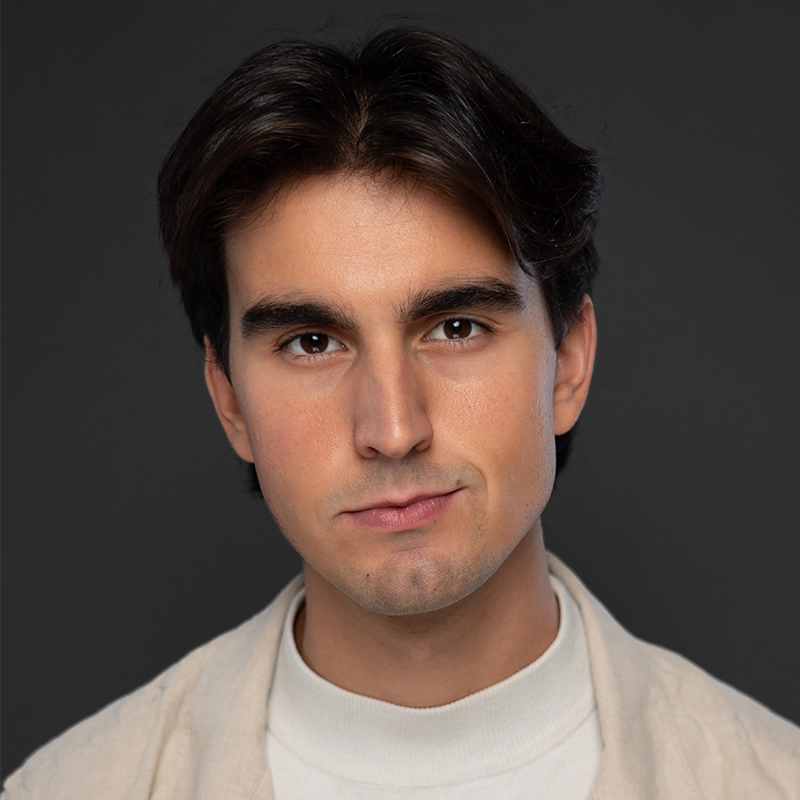 A young man with medium-length dark hair and thick eyebrows looks directly at the camera with a neutral expression. Styled by Katie Yeomans, he wears a light beige jacket over a white turtleneck against a solid dark gray background.
